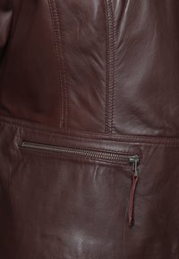 Brun læderjakke sektion med synlige sømme. Har en lynlåslomme med metalhardware og en stoftrækflap langs sømmen.