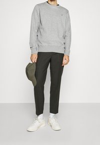 Sudadera gris con puños de canalé, combinada con pantalones verdes oscuros y zapatillas blancas. Se sostiene una gorra verde con la mano derecha.