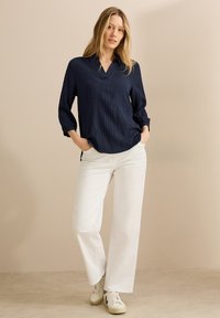 Mariene textuur blouse met een V-hals en driekwart mouwen, gecombineerd met witte wijde broek en lichtgekleurde sneakers.