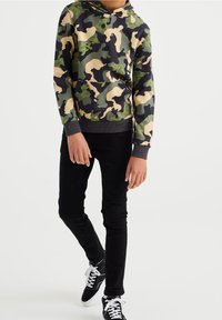 Bambino che indossa un hoodie mimetico verde, beige e nero, pantaloni neri e sneakers alte nere con lacci bianchi, su uno sfondo bianco.