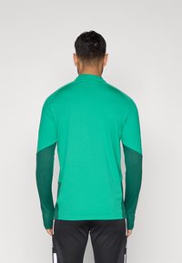 Hosszúujjú sportpóló élénk zöld színben, sima testfelülettel és sötétzöld, texturált ujjakkal. Kényelemre és mozgásra lett tervezve.