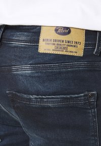 Jeans de mezclilla azul oscuro con una etiqueta de cuero que presenta "Petrol" y texto. Costuras visibles, con bolsillos traseros y un ligero desgaste en la tela.