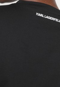 Černé bavlněné tričko s kulatým výstřihem a bílým textem "KARL LAGERFELD" vyšitým na zadní straně blízko výstřihu.
