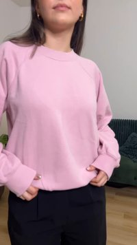 Vaaleanpunainen collegepaita resorilla varustetulla pääntiellä ja hihojen resoreilla, käsien ollessa taskuissa. Yhdistetty tummiin housuihin, neutraalia taustaa vasten.