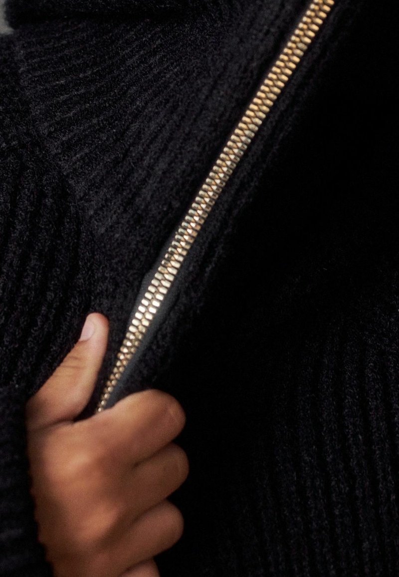 Main tenant un tissu noir tricoté près d'une fermeture éclair métallique dorée partiellement fermée.