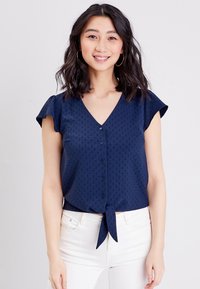 Marineblaue Bluse mit V-Ausschnitt, kurzen schwingenden Ärmeln und einer Bindegarnitur vorne. Sie hat ein strukturiertes Muster und einen Knopfverschluss.