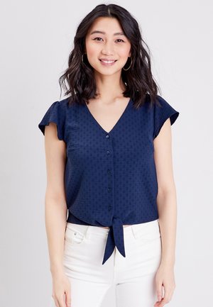 Navyblauwe blouse met een V-hals, korte fladdermouwen en een strikdetail aan de voorkant. Bevat een gestructureerd patroon en knoopsluiting.