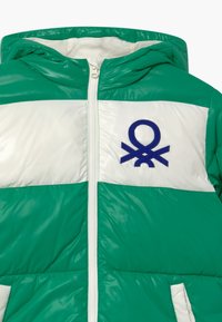 Puffig grön och vit jacka med huva, med dragkedja fram och en blå broderad logotyp på bröstet. Slät, glänsande yta.