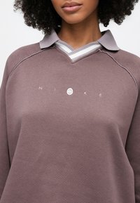 Nike πουλόβερ σε μοβ ύφασμα, με ριμπ γκρι γιακά και σχεδιασμό με λαιμόκοψη σε σχήμα V. Το λογότυπο εμφανίζεται κεντρικά με λευκή κεντητή κεντρική διακόσμηση.