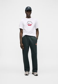 Άνδρας που φοράει λευκό μπλουζάκι Nike, σκούρο παντελόνι φόρμας με ταμπέλα Nike, γκρι αθλητικά παπούτσια και μαύρο καπέλο Nike, στέκεται μπροστά σε απλό λευκό φόντο.