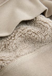 Beige Kleidungsstück mit einer gerippten Außenschicht und einem weichen, flauschigen Innenfutter. Die Nähte sind entlang der Kanten sichtbar, was den Texturkontrast verstärkt.