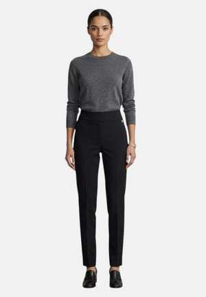 Donna in piedi che indossa un maglione grigio a maniche lunghe, pantaloni sartoriali neri a vita alta, mocassini neri e un braccialetto d'oro al polso destro.