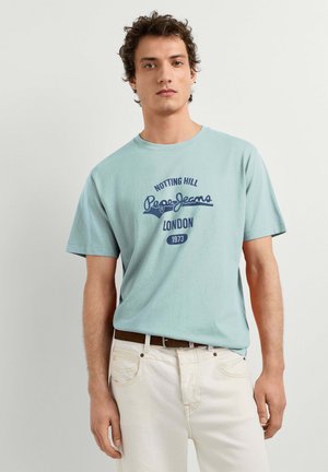 Młody mężczyzna z kręconymi włosami, ubrany w jasnoniebieską koszulkę Pepe Jeans London i białe spodnie, stojący na tle jednolitego jasnego tła.