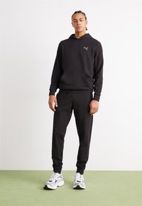 Il felpa nera presenta una tasca a marsupio e polsini a coste, abbinata a pantaloni jogger neri e sneakers atletiche bianche.