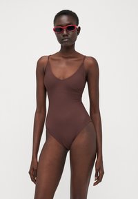 Maillot de bain une pièce brun à côtes avec un décolleté en V, des fines bretelles et un design à jambes hautes. Le modèle porte des lunettes de soleil rouges.