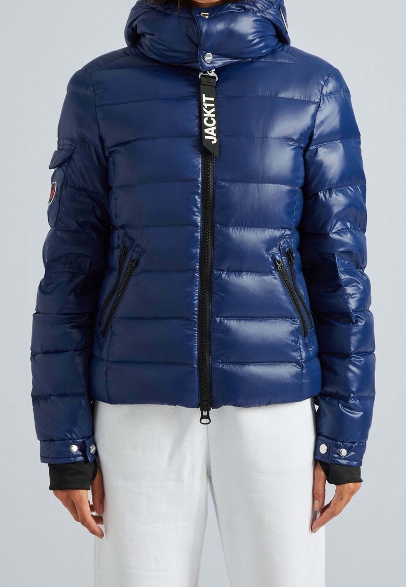 Personne portant une doudoune bleu marine brillante avec capuche, fermeture éclair à l'avant, poches zippées, et un pantalon blanc, debout devant un fond uni.