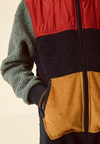 Giacca in pile con parte superiore trapuntata rossa, maniche testurizzate verdi, corpo blu navy e tasche in corduroy arancioni. Presenta una zip frontale e polsini a coste.