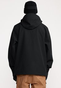 Veste noire imperméable avec capuche, présentant une coupe décontractée et une texture lisse. Portée sur un pantalon beige, vue de dos.