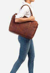 Bolso tote de cuero marrón con acabado texturizado, que presenta asas dobles y cierre de cremallera, llevado por una persona en jeans ajustados.