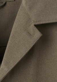 Blazer verde oliva con un tejido texturizado, cuello con muescas afiladas y un detalle de costura mínima en la solapa y el área del bolsillo.