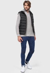 Mænds sort puffervest over en lys grå striktrøje, blå tætsiddende jeans med paneldetaljer og hvide sneakers med sort sål.