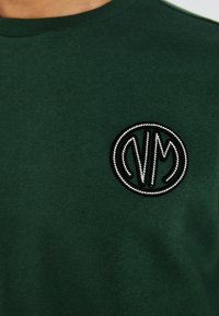 T-shirt verde in cotone con un logo ricamato nero rotondo e dettagli bianchi, realizzato su un tessuto liscio e opaco.