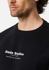 Svart sweatshirt i bomullsblandning med rund halsringning. Framsidan visar vit logotyptext som inkluderar "Slade Studios" och en tagline.