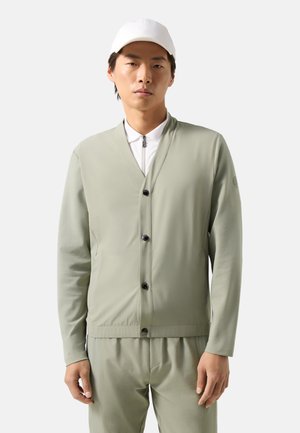 Młody mężczyzna ubrany w blady zielony sweter zapinany na guziki, dopasowane spodnie, białą koszulę zapinaną na zamek i białą czapkę z daszkiem, stojący na białym tle.