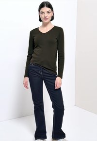 Olivgrünes, langärmliges V-Ausschnitt-Top kombiniert mit dunkelblauen, ausgestellten Jeans mit heller Naht und figurbetonter Silhouette.