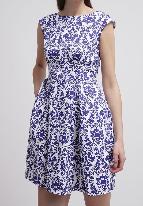 Femme portant une robe blanche sans manches avec un motif floral damassé bleu, taille cintrée et jupe plissée, debout avec une main dans la poche.