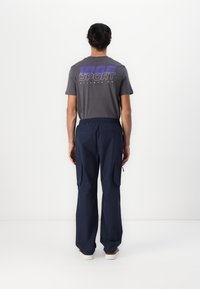 Homme debout face à un mur blanc, portant un t-shirt gris avec un texte bleu, un pantalon cargo bleu marine avec des poches et des chaussures blanches.