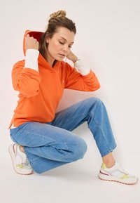 Oranger Kapuzenpullover mit weißen Bündchen, kombiniert mit hellblauen weitem Jeans und weißen Sneakers mit Pastellakzenten.