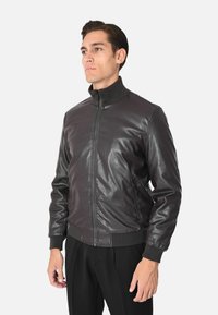 Giacca bomber in pelle marrone scuro con colletto alto, chiusura frontale con cerniera e polsini e orlo a costine. Presenta tasche laterali e una texture liscia.