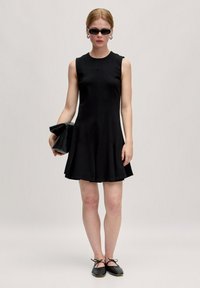 Zwarte mouwloze jurk met een uitlopende zoom, gemaakt van een gladde stof. Model houdt een zwarte clutch vast, draagt kleine zwarte zonnebrillen en platte schoenen.