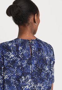 Top azul com padrão que apresenta um design texturizado, decote redondo, mangas curtas e um detalhe em forma de olho-chave nas costas, preso com botões dourados.