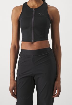 Nainen, jolla on musta hihaton vetoketjullinen crop top ja mustat housut, seisoo yksiväristä taustaa vasten.