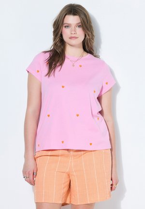 Junge Frau trägt ein pinkes T-Shirt mit kleinen orangen Herzen und pfirsichfarbene gestreifte Shorts und steht vor einem weißen Hintergrund.