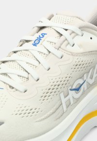 Close-up de um sapatilha de corrida Hoka bege com atacadores brancos, parte superior em malha respirável e um detalhe amarelo na sola.