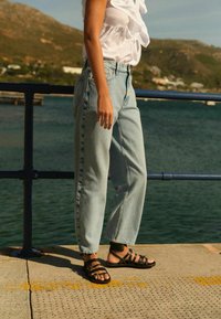 svetlobluje visoke jeans hlače, sproščen kroj, z zbledelim izgledom in ravnimi nogavicami. Črne vezane sandale, ki razkrivajo prste. Bele bluze z naborki.