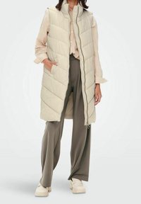 Jasno beżowa kurtka bez rękawów z wysokim kołnierzem, skośnym pikowaniem i bocznymi kieszeniami, zestawiona z jasną koszulą i szerokimi szarymi spodniami.