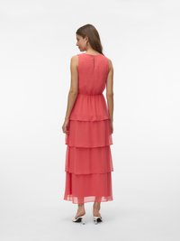 Femme vue de dos portant une robe maxi corail rose sans manches à volants avec un motif subtil de points et des sandales blanches à talons, sur fond blanc.
