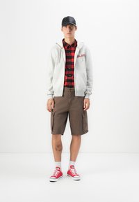 Giovane uomo con cappellino nero, felpa grigia con zip, camicia a quadri rossa e nera, pantaloni cargo marroni, calze bianche e scarpe da ginnastica rosse, in piedi.
