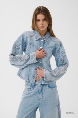 Femme portant une veste en jean brodée bleu clair et un jean assorti sur un fond uni, ajustant les boutons de la veste.