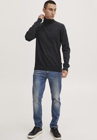 Pulover turtleneck de culoare gri închis cu un model texturat, asortat cu blugi din denim bleu deschis și pantofi sport negri. Modelul stă cu mâna pe gât.