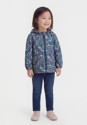 Niña joven sonriendo, con chaqueta con capucha de flores, jeans azules y zapatos rosados claros, de pie contra un fondo blanco.