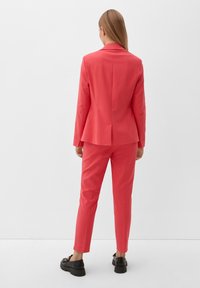 Costume corail avec une veste ajustée et un pantalon fuselé. Tissu lisse, deux fentes au dos de la veste, associé à des chaussures noires.
