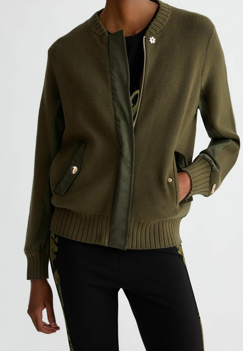 Personne portant une veste bomber en maille vert olive avec des accents rembourrés, des boutons dorés et un pantalon noir avec des rayures vertes à motifs.