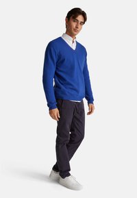 Blauer V-Ausschnitt-Pullover aus weichem Material, kombiniert mit dunkler Hose und weißen Sneakers. Das Outfit vereint lässige und entspannte Designelemente.