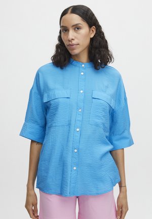 Chemise bleue à manches courtes avec une finition texturée, deux poches avant et un col mandarin. Comprend des boutons blancs et une coupe décontractée.