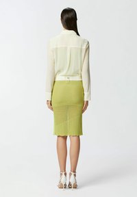 Blusa leggera beige chiaro e trasparente con colletto, abbinata a una gonna in maglia verde brillante lunga fino al ginocchio con orlo a onde. Sandali bianchi con tacco.